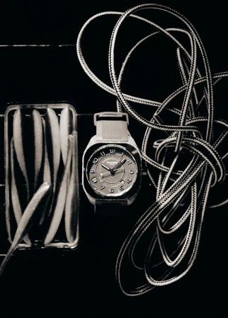 Still life photo of a wristwatch next to a clear container filled with oval shapes, and rope against a dark background.