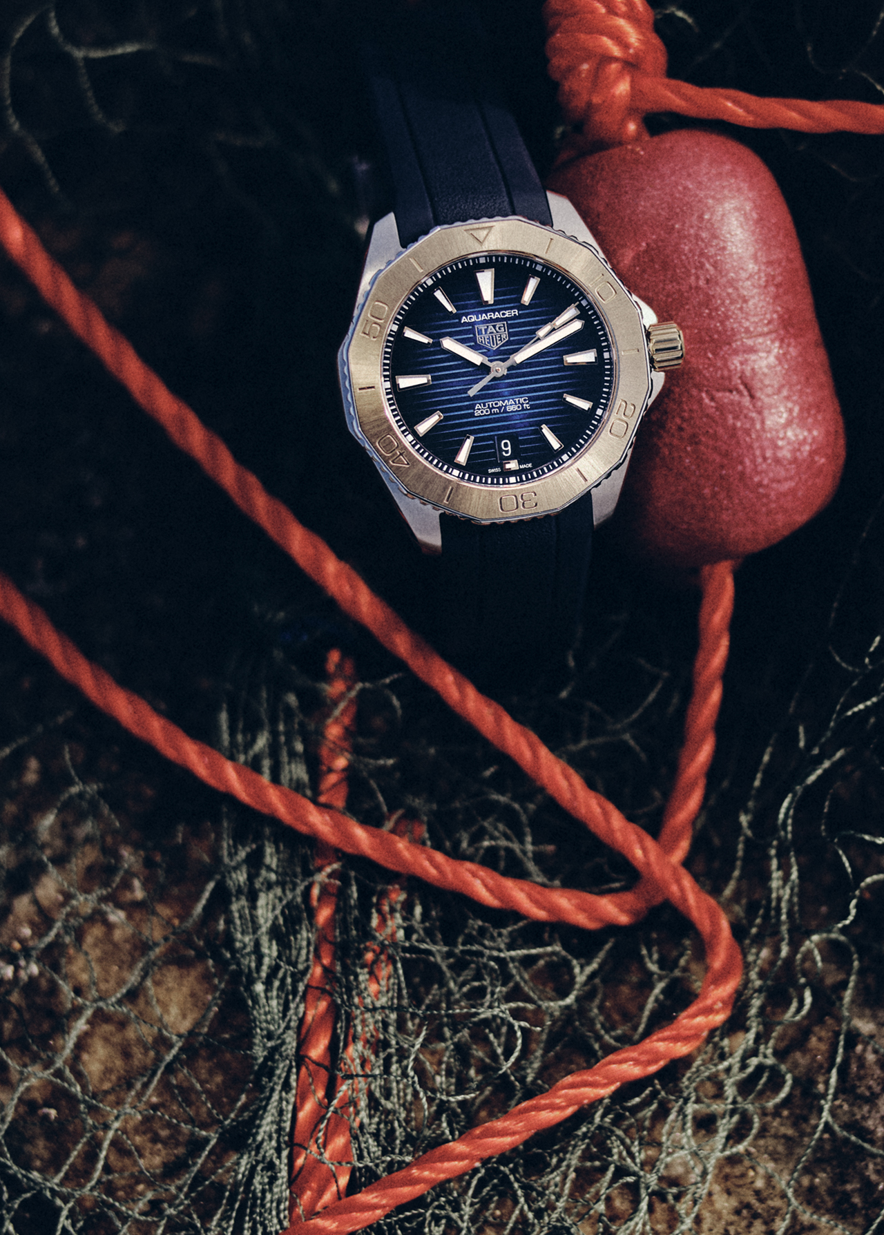 Wristwatch with a blue dial and gold bezel is displayed with an orange rope and fishing net, set against a dark background.