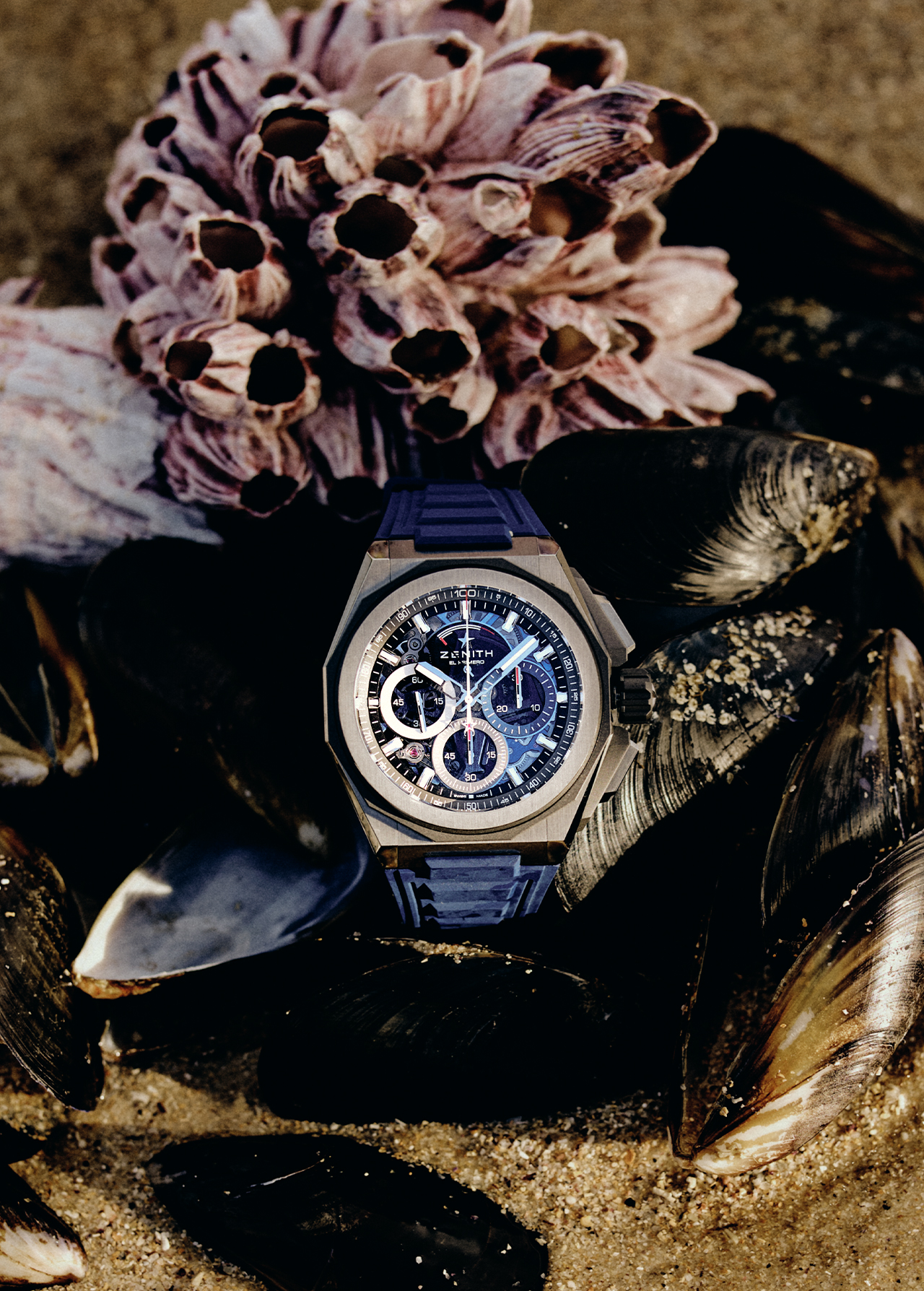 Zenith Defy watch surrounded by shells and barnacles on wet sand, close-up studio shot, highlighting luxurious design and beachy vibe.