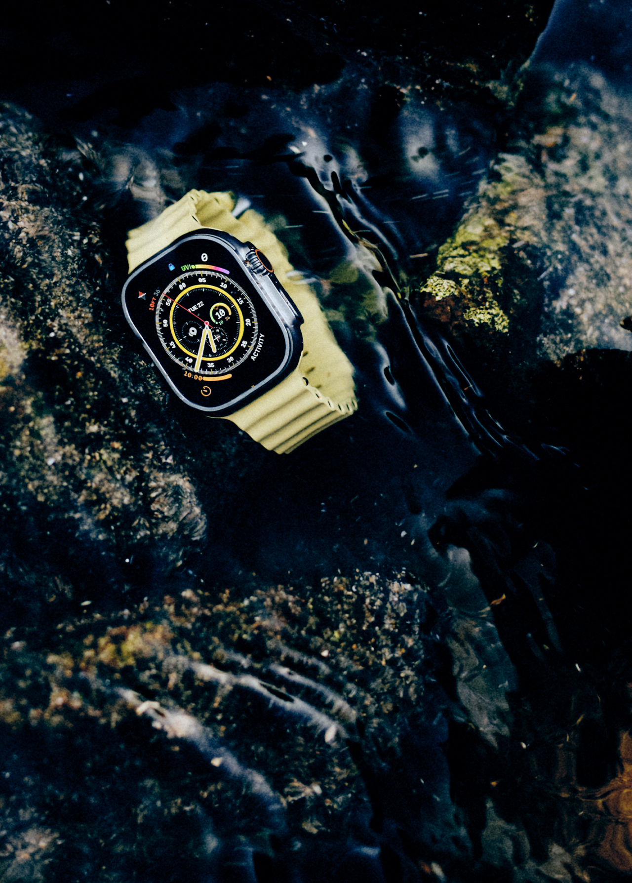 Close-up of a smart watch with a yellow wristband submerged in shallow water with a rocky bottom for outdoor activities.