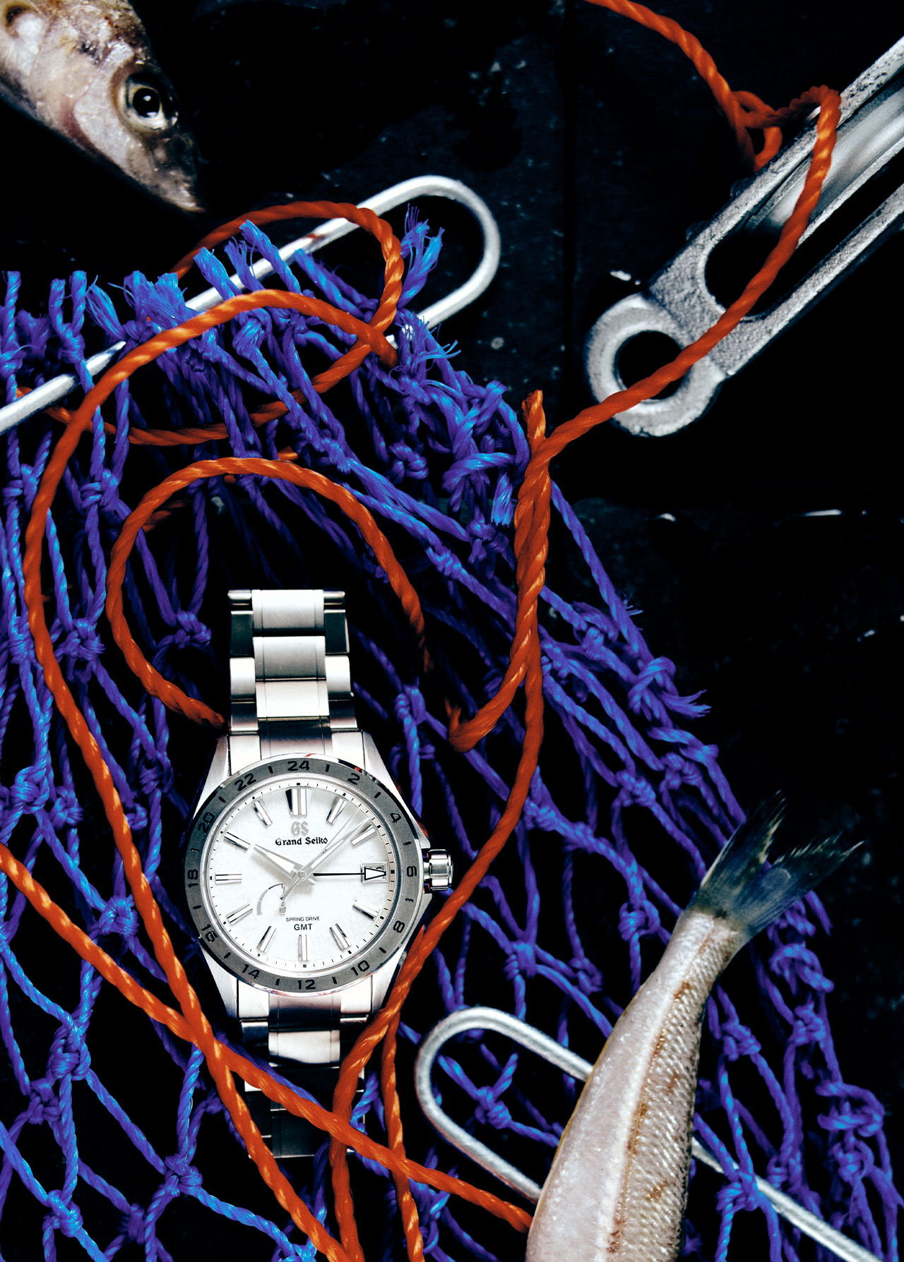 Luxury wristwatch with a silver band and white face displayed with a fishing net, rope, metal hardware, and fish over a dark background.