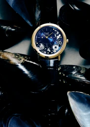 Close-up product photograph of a luxury gold watch with a deep blue dial surrounded by dark glossy mussel shells.
