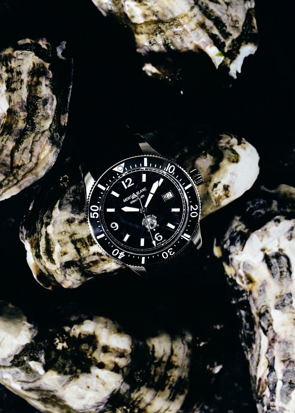 Close-up product shot of a luxury dive watch with a black dial surrounded by opened oysters on a dark surface