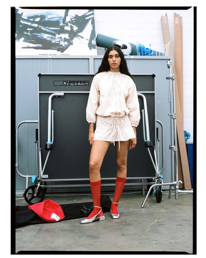 Woman poses in a light jacket and skirt in front of a ping pong table in an indoor studio setting with concrete floors.