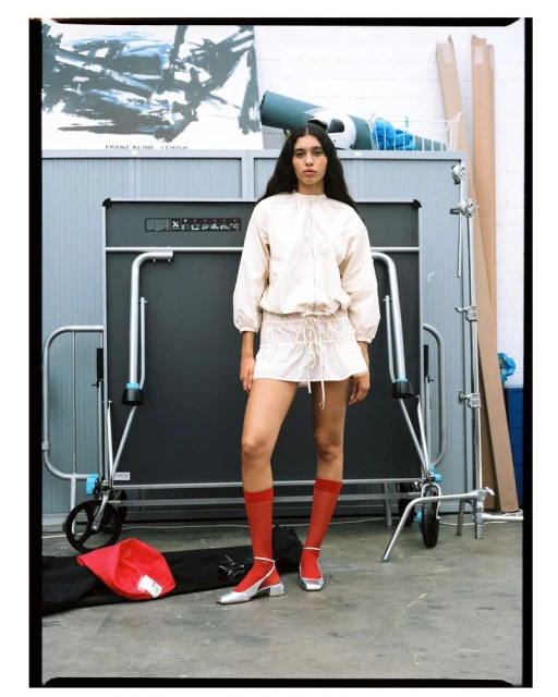 Woman poses in a light jacket and skirt in front of a ping pong table in an indoor studio setting with concrete floors.