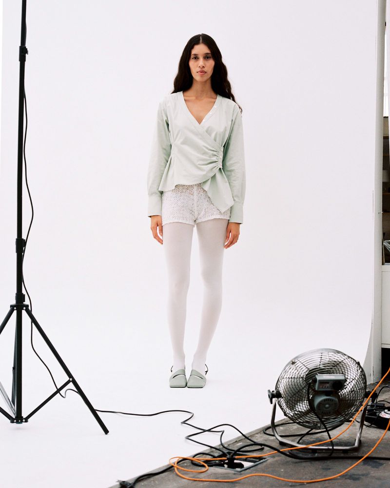 Full-length shot of a woman in light green top, white shorts and tights, standing in a photo studio with lighting and fan equipment.