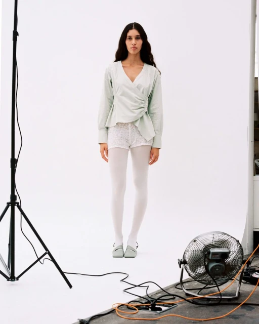 Full-length shot of a woman in light green top, white shorts and tights, standing in a photo studio with lighting and fan equipment.