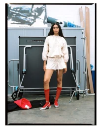 Model in cream skirt set with red socks and silver shoes standing in a studio with equipment.