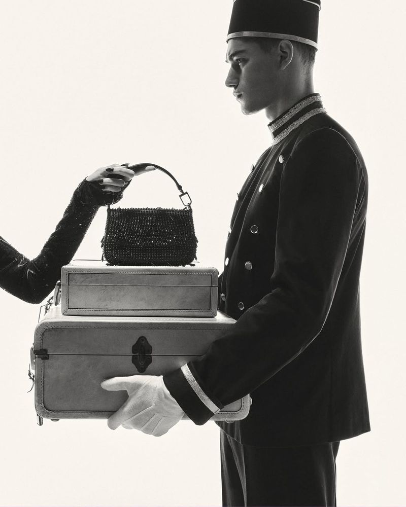 A monochromatic shot presents a bellhop holding stacked luggage as a hand reaches for a purse resting on top of the suitcases.