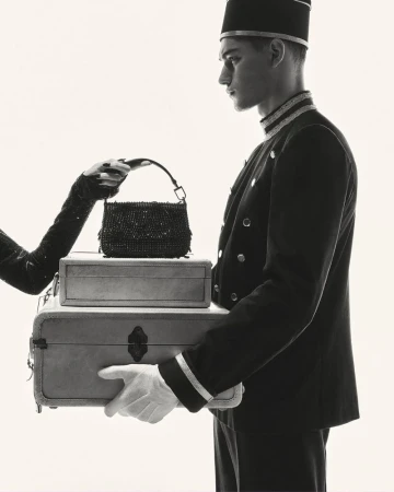 A monochromatic shot presents a bellhop holding stacked luggage as a hand reaches for a purse resting on top of the suitcases.