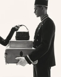 Profile of a male bellhop in uniform holding stacked luggage while a gloved hand places a beaded handbag on top against a bright white backdrop