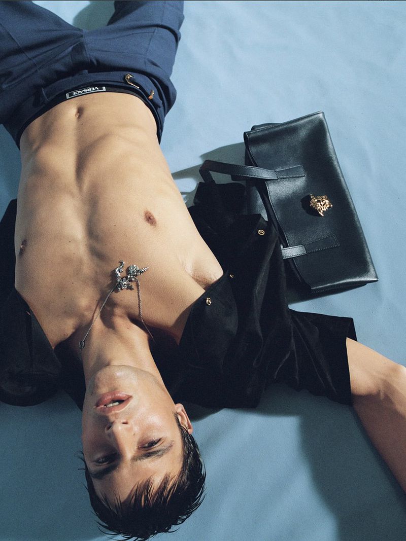 Male fashion model poses shirtless with navy suit trousers and black leather bag in studio shot with light blue backdrop.