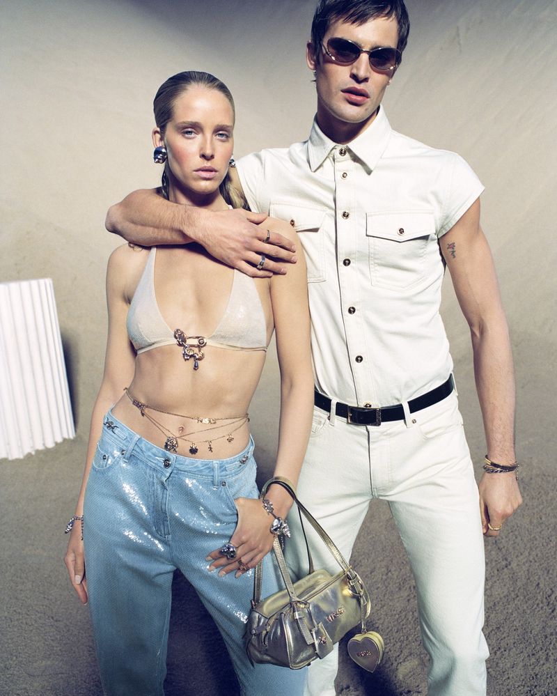 Two fashion models in a beige indoor studio setting. A woman in a bralette and jeans stands beside a man in a button-down shirt and pants.