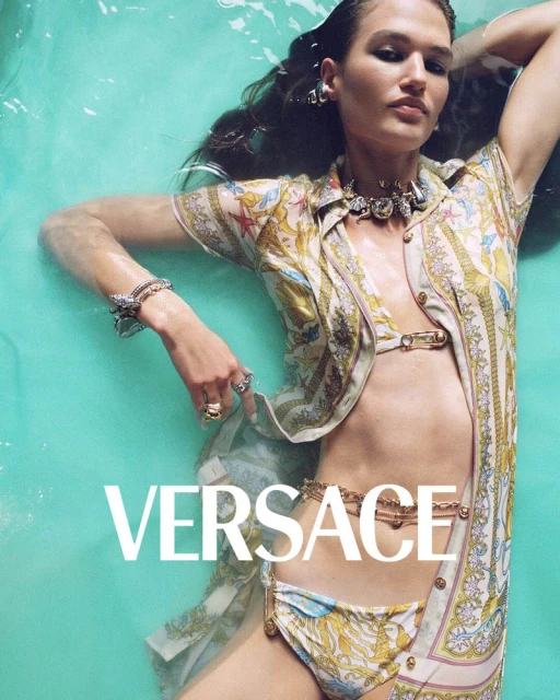 A woman with long dark hair poses in turquoise water wearing a patterned top, bikini bottom, and jewelry in a fashion shot.