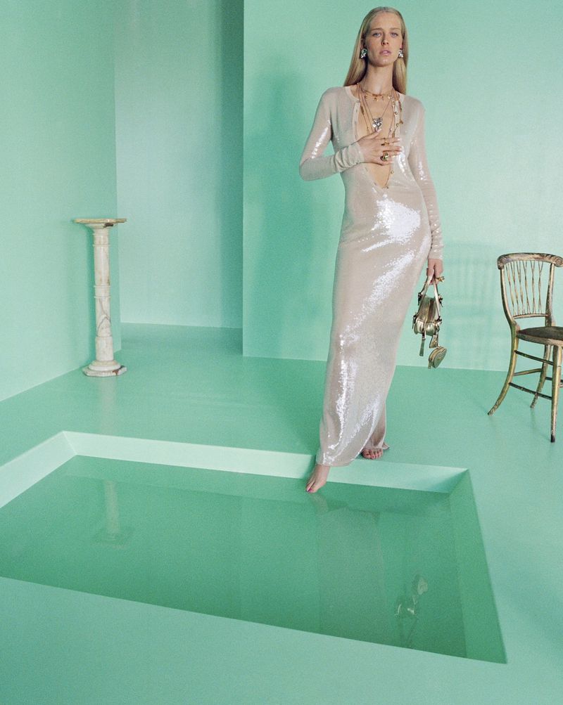 A blonde woman in a champagne-colored sequin gown stands in a room with turquoise floors and walls, stepping into a pool of water.