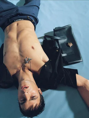 Shirtless male model wearing blue trousers and a black shirt lying on a light blue background beside a black handbag.