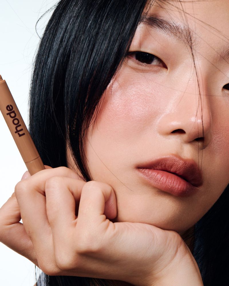 Close-up of an Asian woman with black hair holding a Rhode brand lip product against a white backdrop for a beauty advertisement.