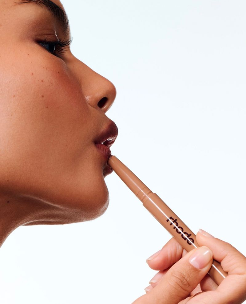 Close-up of a person applying a nude lip liner to their upper lip. The person has tan skin and is positioned in profile.