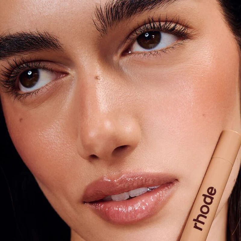 Close-up studio shot of a woman with brown eyes, wearing a nude lipgloss, and holding a skincare product, Rhode, in a vertical format.