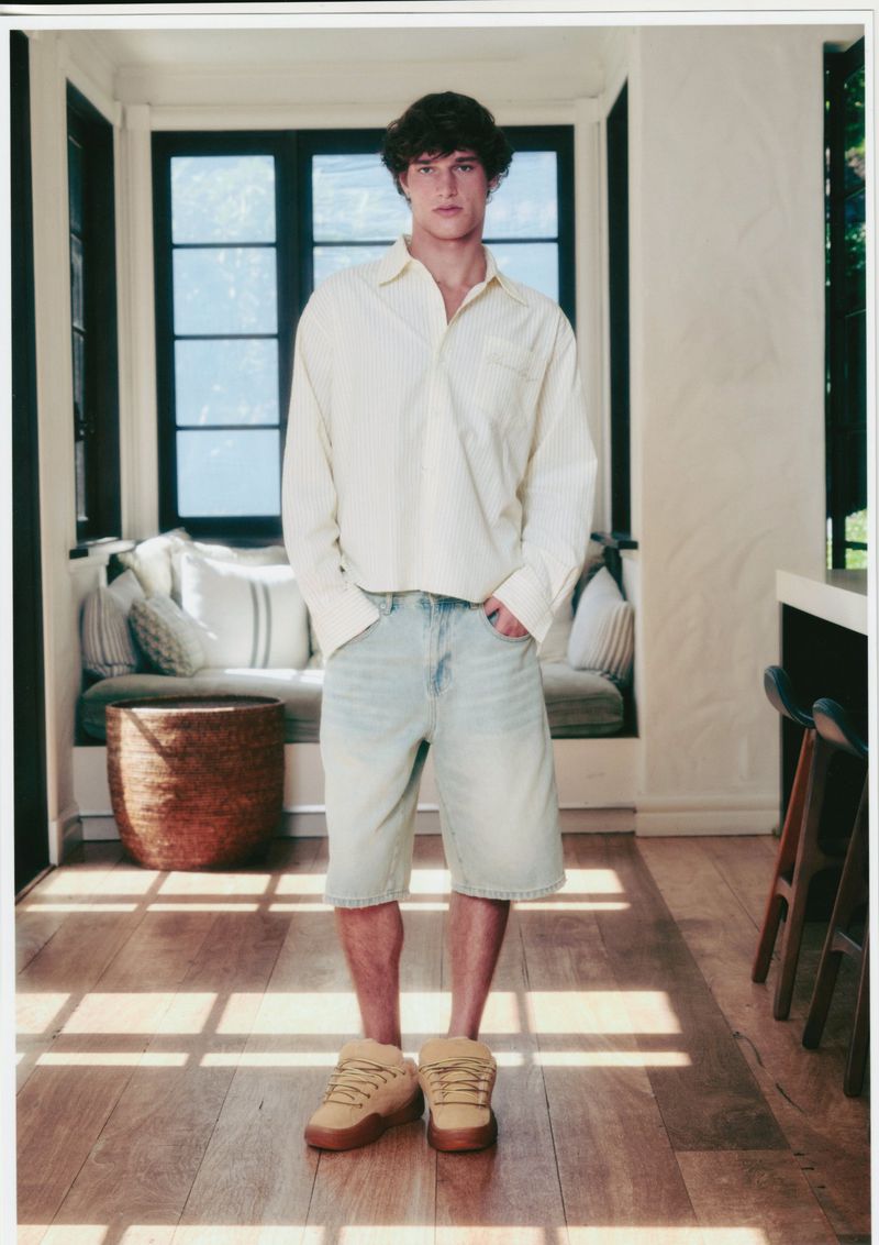 An adult male stands indoors wearing a button-up shirt, denim shorts, and platform shoes, illuminated by bright, natural light.