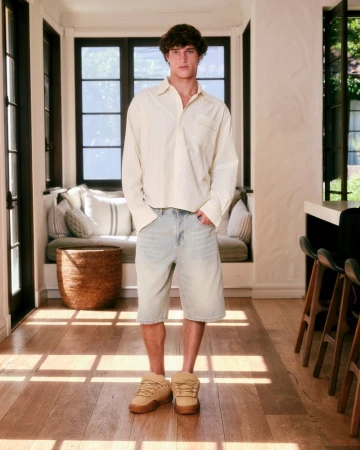 A young adult male stands in a home interior, wearing a yellow striped shirt, light blue jean shorts, and tan sneakers in full shot.