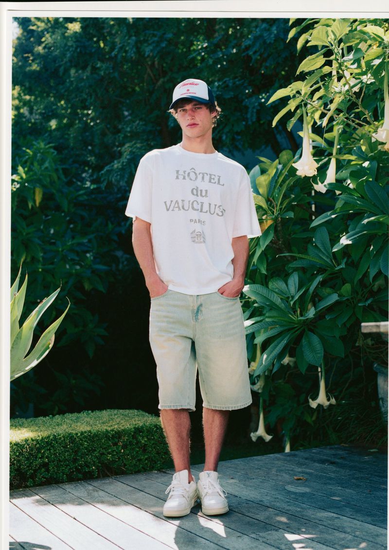 Young adult male standing outdoors wearing a white graphic t-shirt, light blue denim shorts, baseball cap, and sneakers. Natural light.