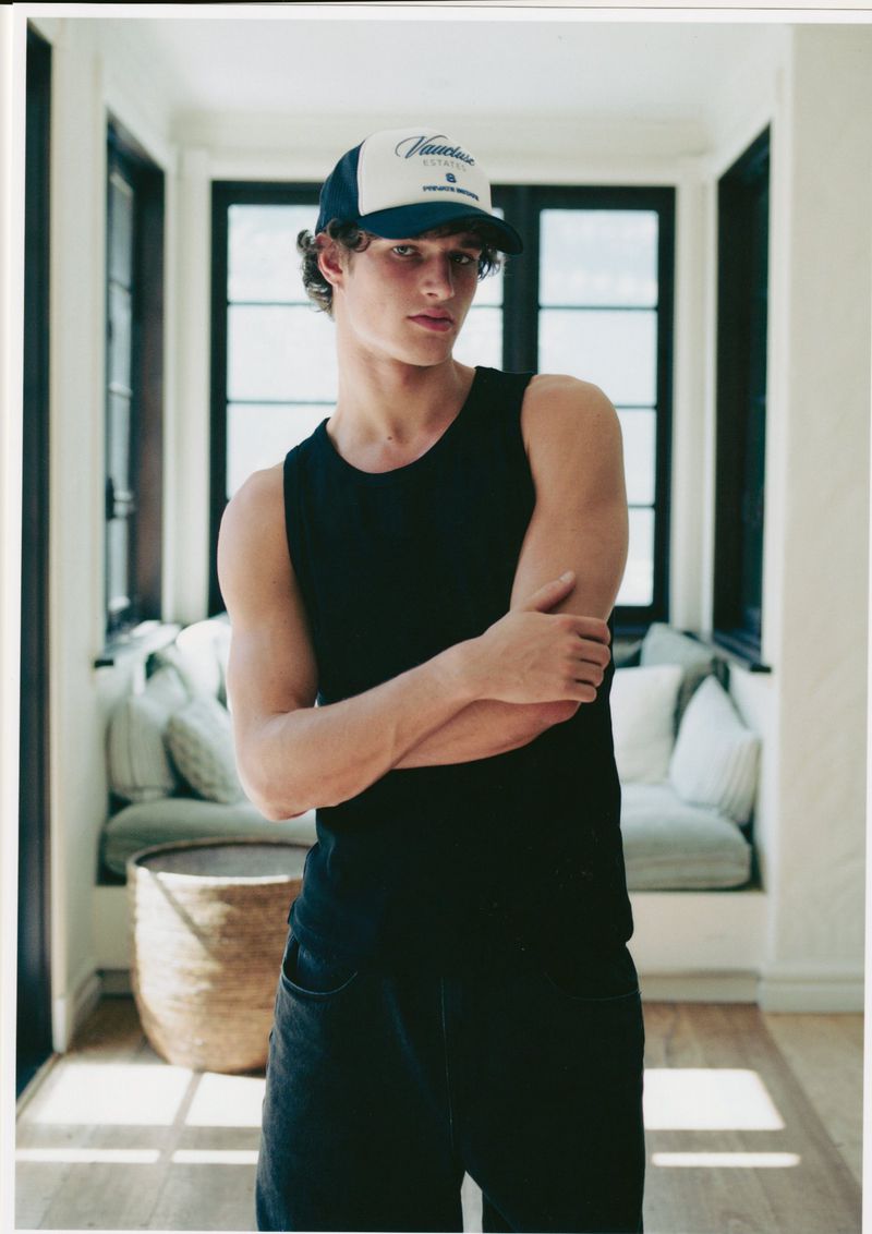 Young adult male in tank top and baseball cap poses indoors with arms crossed against a neutral backdrop with natural lighting.