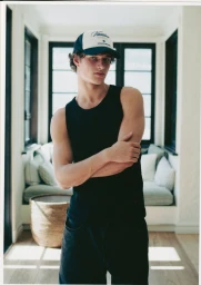 Model wearing a black tank top and trucker hat standing in an interior space with a window and daybed in the background.