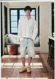 A young male model wearing an ivory long-sleeve shirt and light denim shorts standing indoors in a bright room.