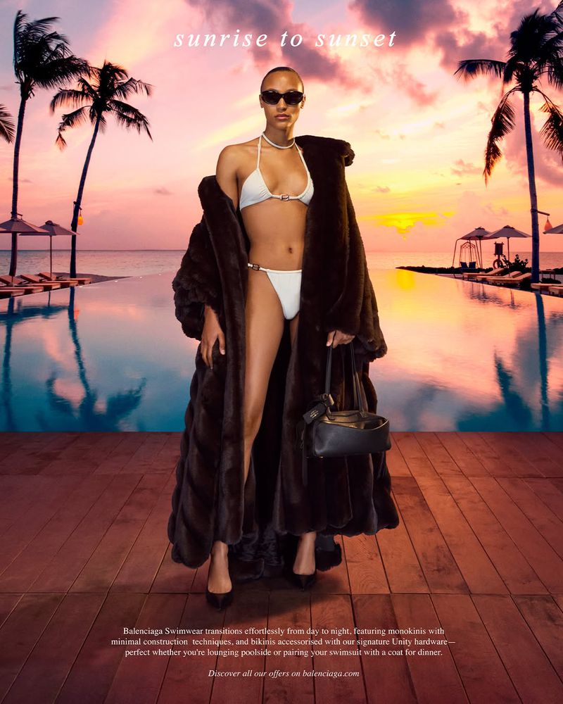 Woman poses in a white bikini and dark coat on a wooden platform with a tropical ocean and sunset background. She also carries a purse.