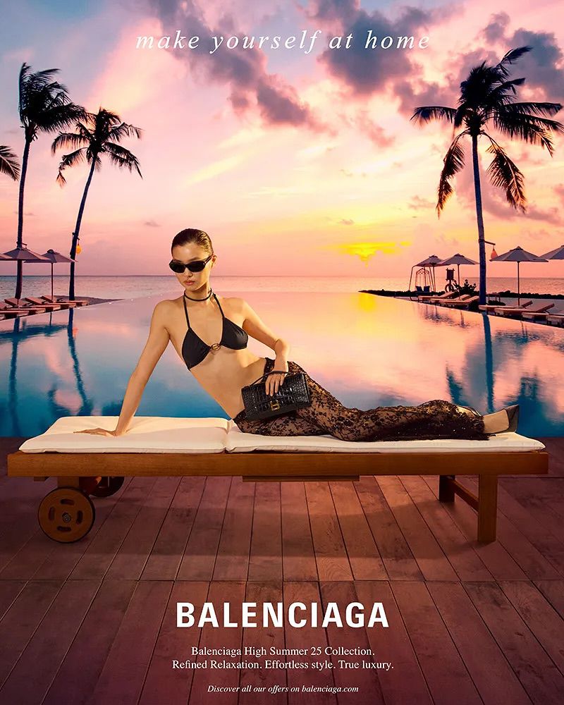 Fashion ad featuring a woman in a black bikini and pants lying on a lounge chair at a tropical resort with a sunset ocean view.