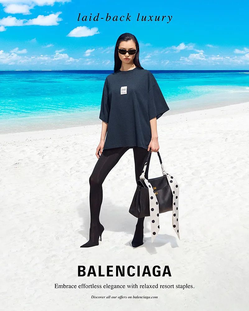 Fashion shot of a woman in sunglasses, black leggings and t-shirt, holding a black purse on a beach with turquoise water.