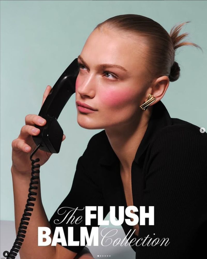 Portrait of a blonde woman holding a black telephone receiver next to her face with makeup and a black shirt against light blue.
