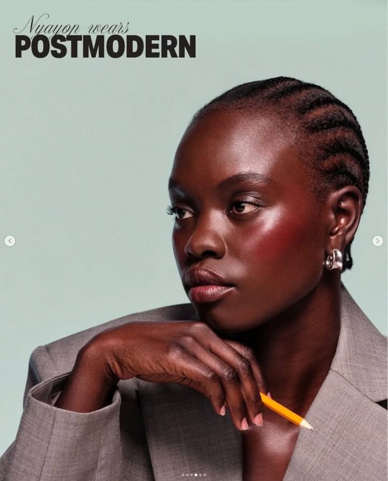 Headshot of a dark-skinned woman with braided hair wearing a grey blazer and holding a yellow pencil against a light-blue background.