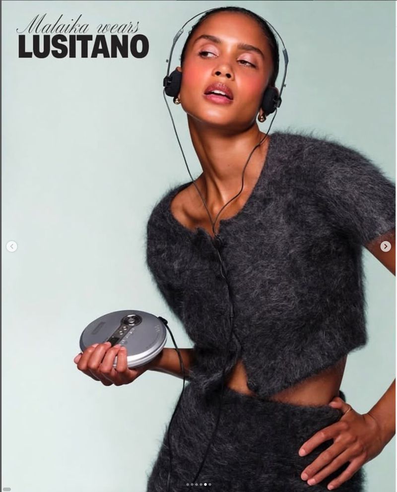 Woman in grey knitwear poses indoors with headphones and retro CD player, lit by soft lighting. Fashion, music, and technology merge.