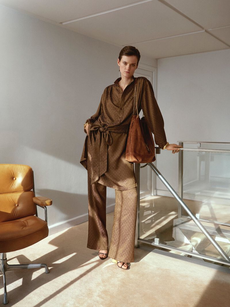 A woman with short hair poses indoors wearing a brown patterned suit with a matching handbag, standing near a metal railing and chair.