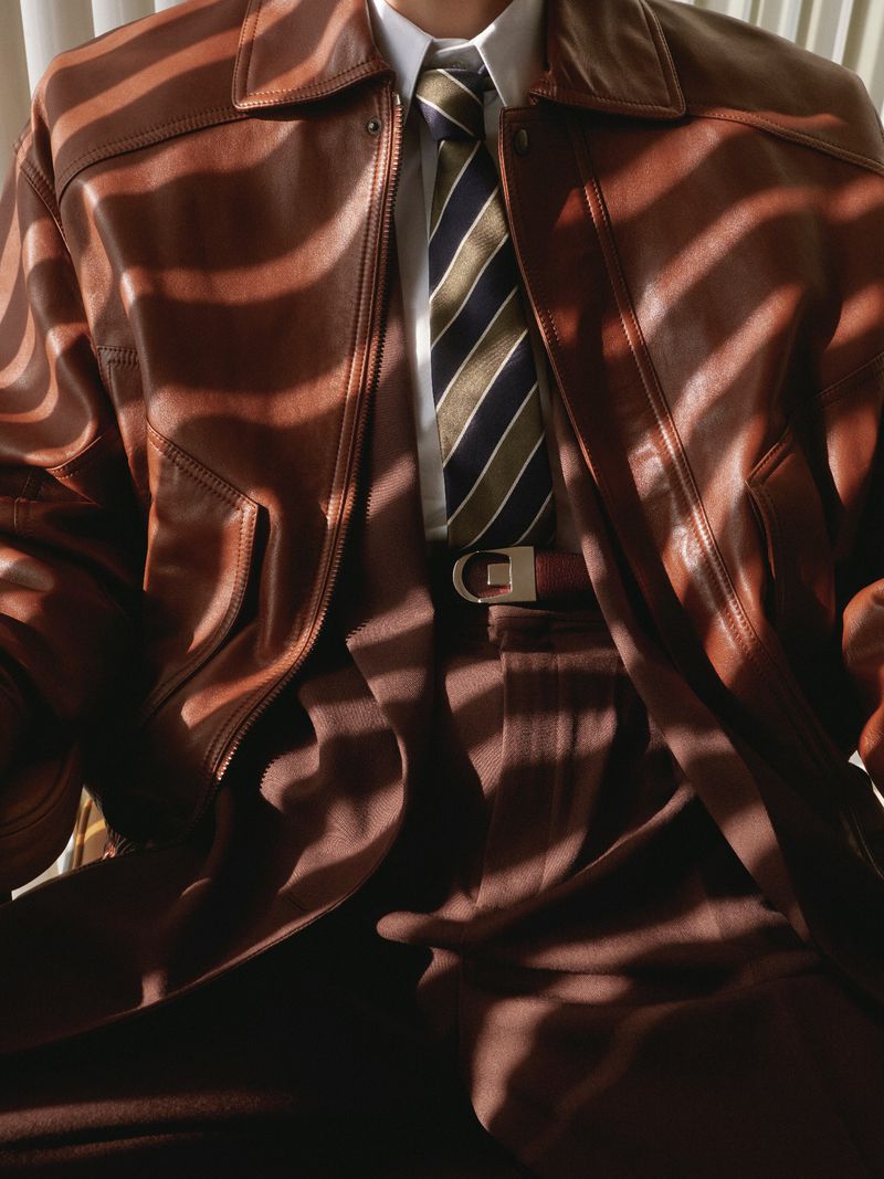Close-up of a person wearing a brown leather jacket, white shirt, striped tie, and brown trousers under striped shadow lighting
