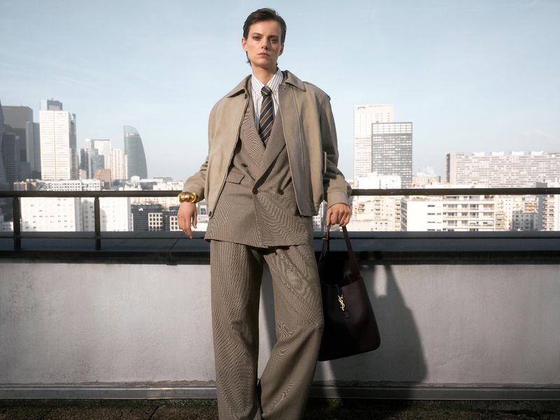 Stylish adult poses on rooftop with city skyline backdrop, wearing suit jacket, trousers, tie, and bomber jacket with Yves Saint Laurent bag.