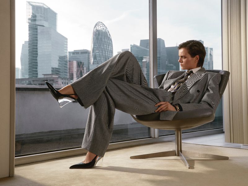 Stylish woman in a grey suit and black heels lounges in a modern chair with a cityscape view from a high-rise window.