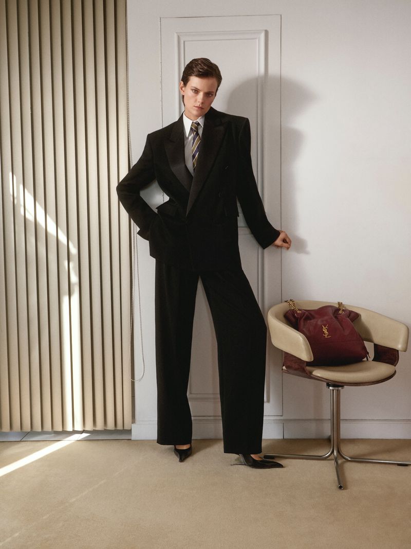 Woman in a black suit standing against a white wall, with a designer handbag on a chair, illuminated by natural light streaming through blinds.
