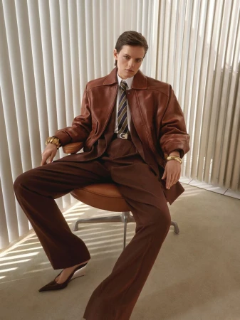 Model seated in leather jacket and brown trousers against vertical blinds for Mytheresa Saint Laurent