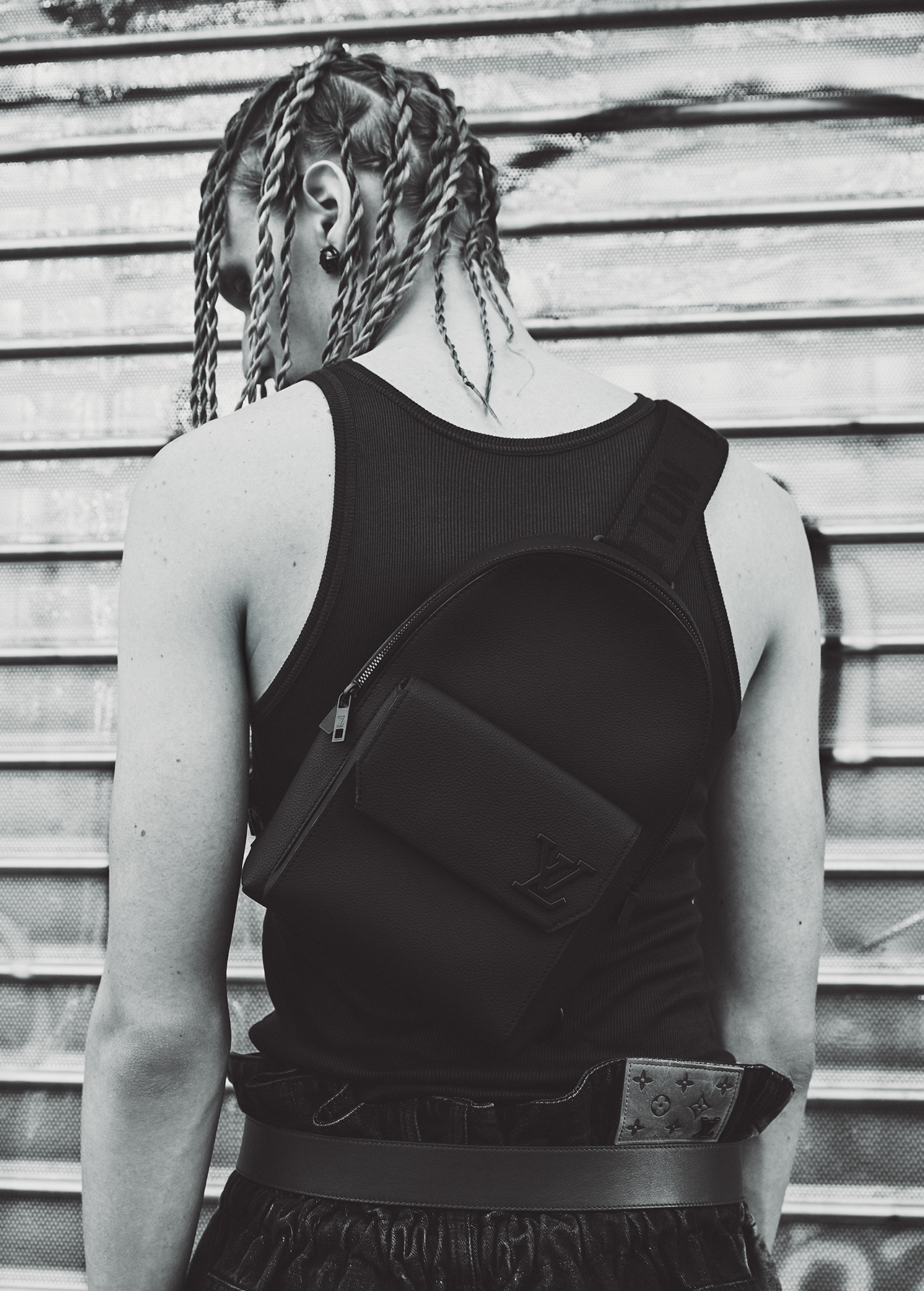 Monochrome portrait of a person with braided hair wearing a black tank top and crossbody bag against a metal shutter door.