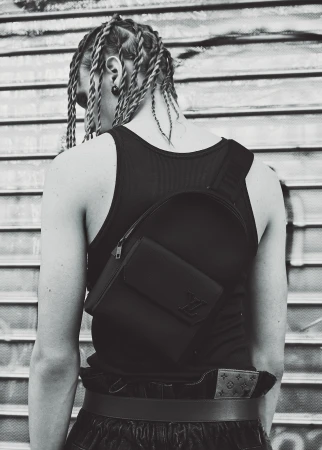 Monochrome portrait of a person with braided hair wearing a black tank top and crossbody bag against a metal shutter door.