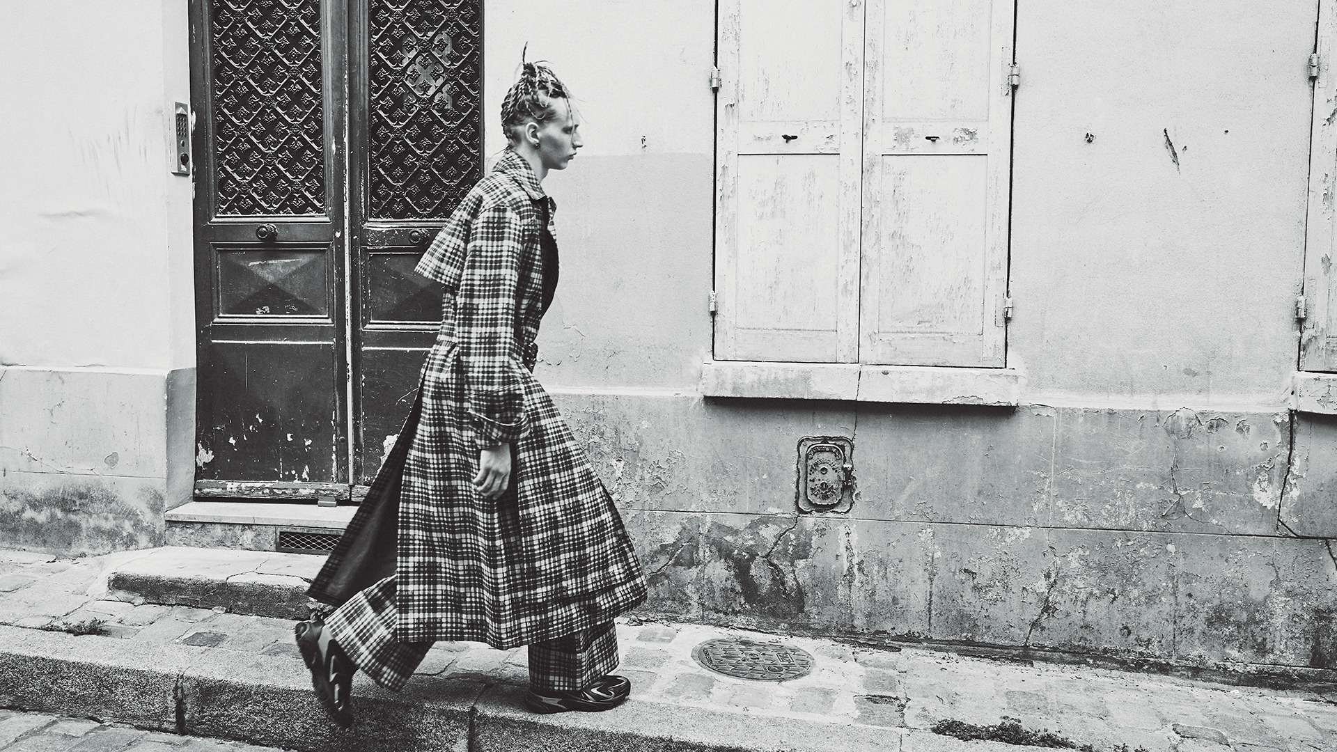 Monochrome fashion shot of a person in a plaid coat walking past a building with old doors and windows in a city street.