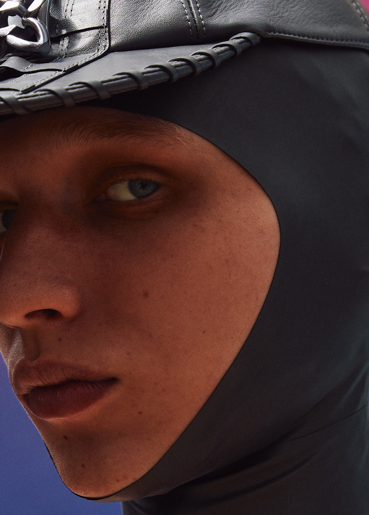 Close-up shot of a fair-skinned person with blue eyes wearing a black leather balaclava and hat against a blurred colorful background.