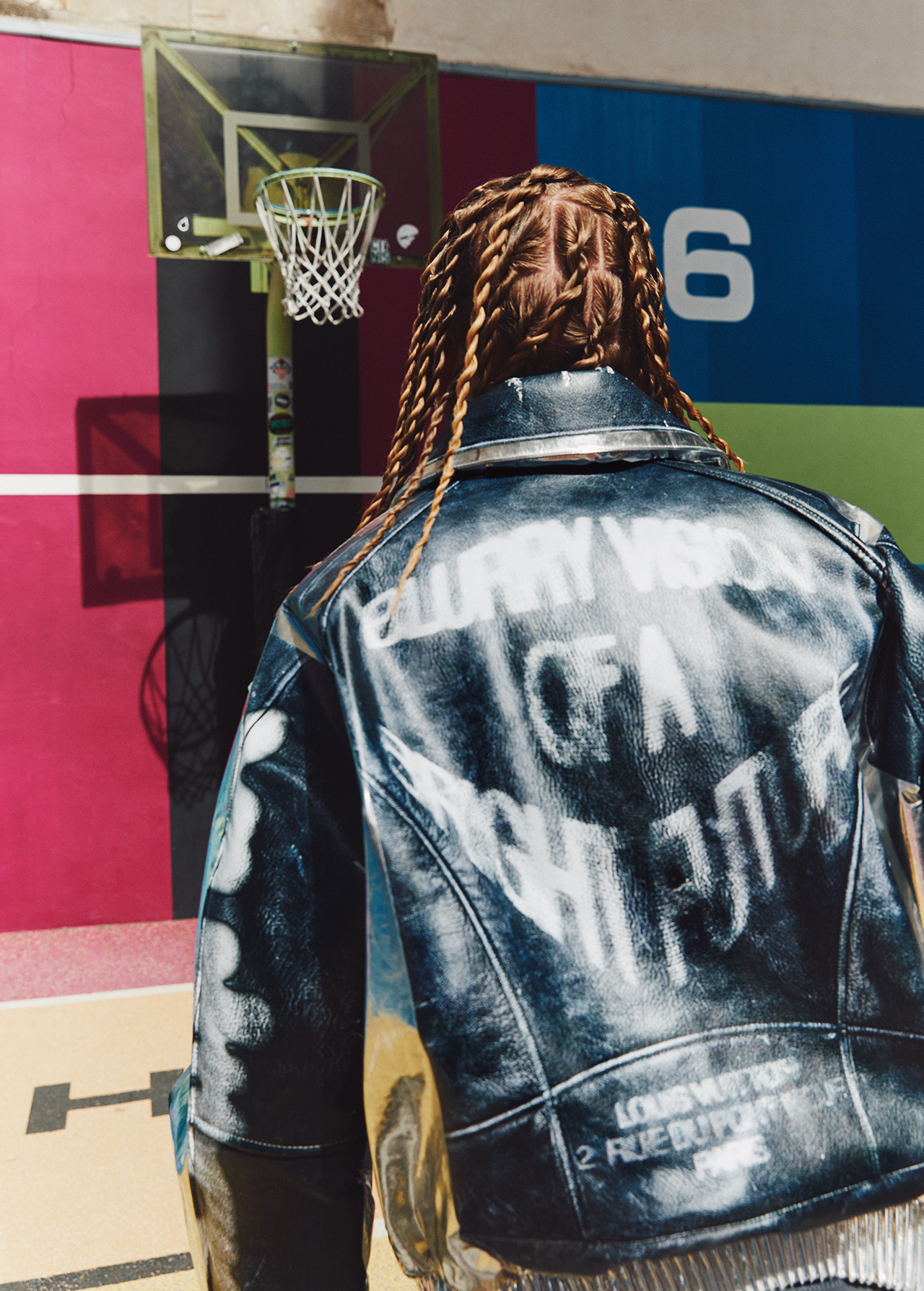 Adult woman wearing black leather jacket with light brown braided hair standing in a basketball court next to a basketball hoop.