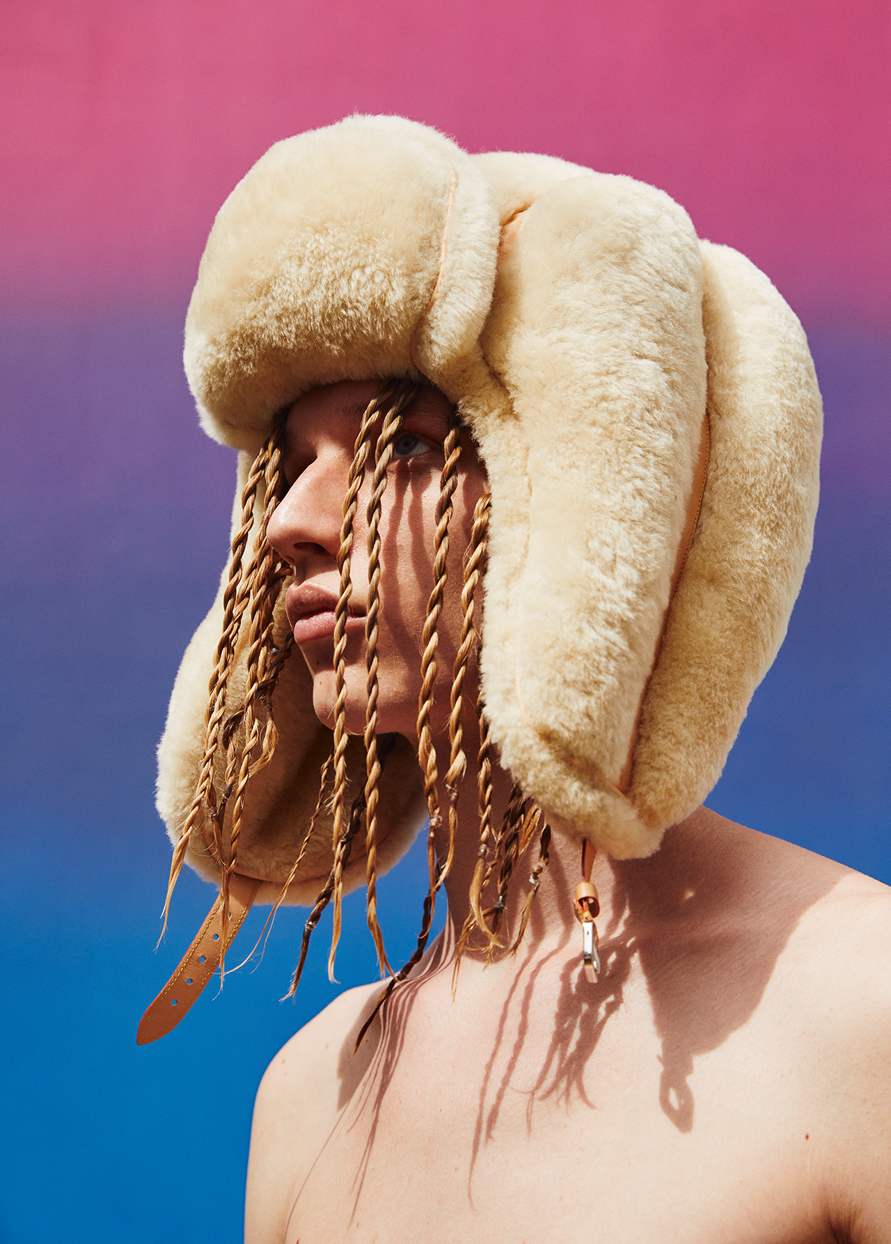 Adult with blonde braids wears a cream-colored fur hat against a background with pink on top and blue on the bottom.