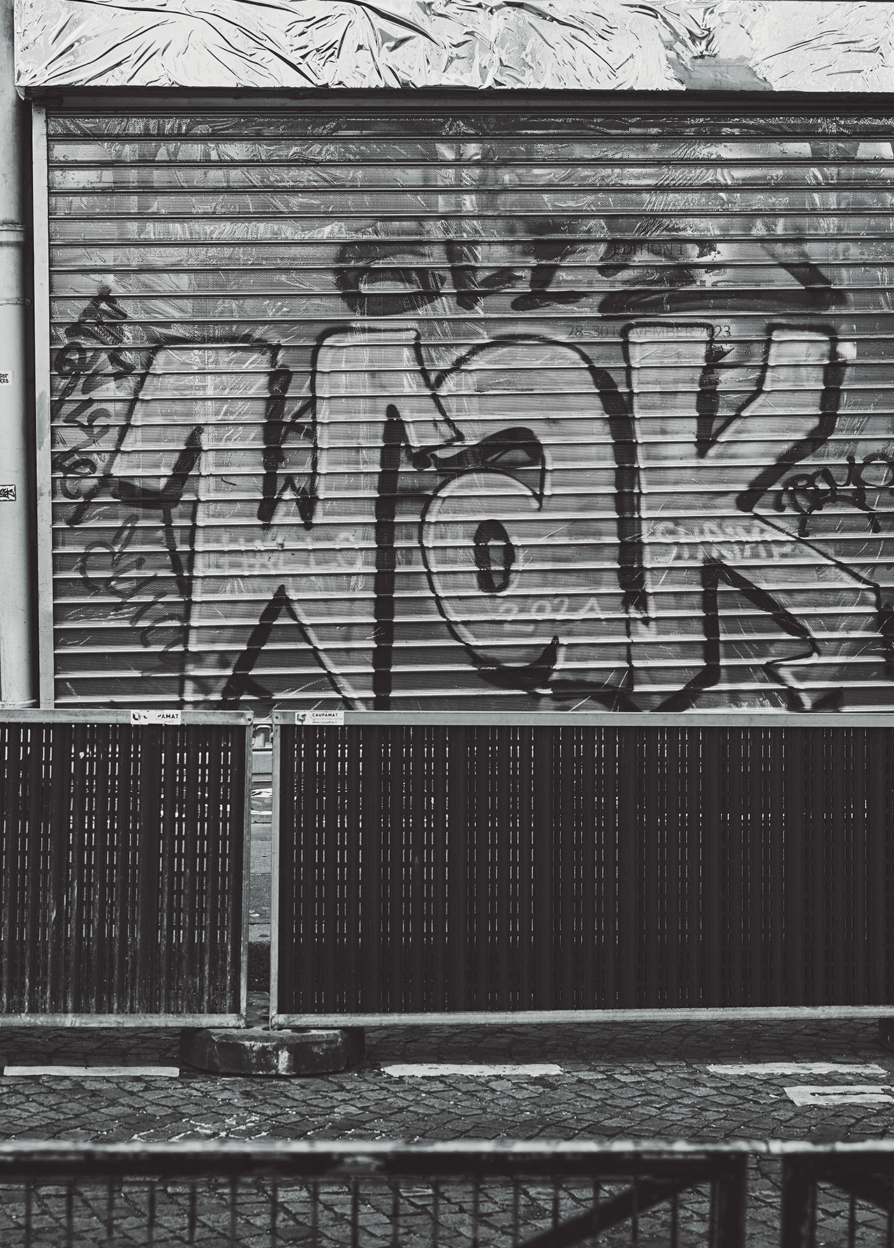 Black and white image features a metal roll-up door covered in graffiti, a fence in front, and textured pavement leading up to the structure.