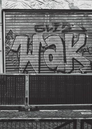 Black and white image features a metal roll-up door covered in graffiti, a fence in front, and textured pavement leading up to the structure.