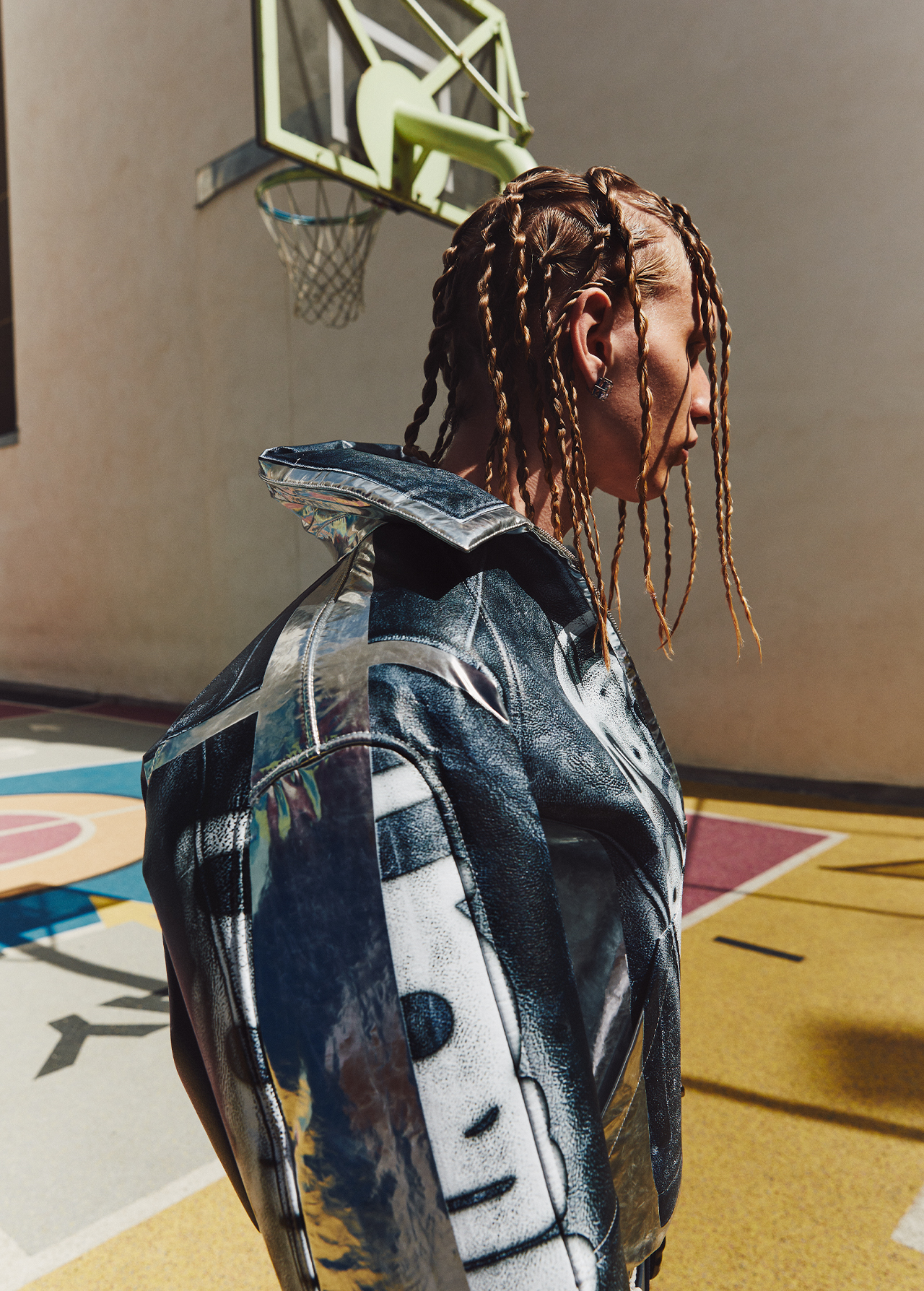 Profile view of a person with blonde braids wearing a shiny silver jacket stands on a yellow and red outdoor basketball court.